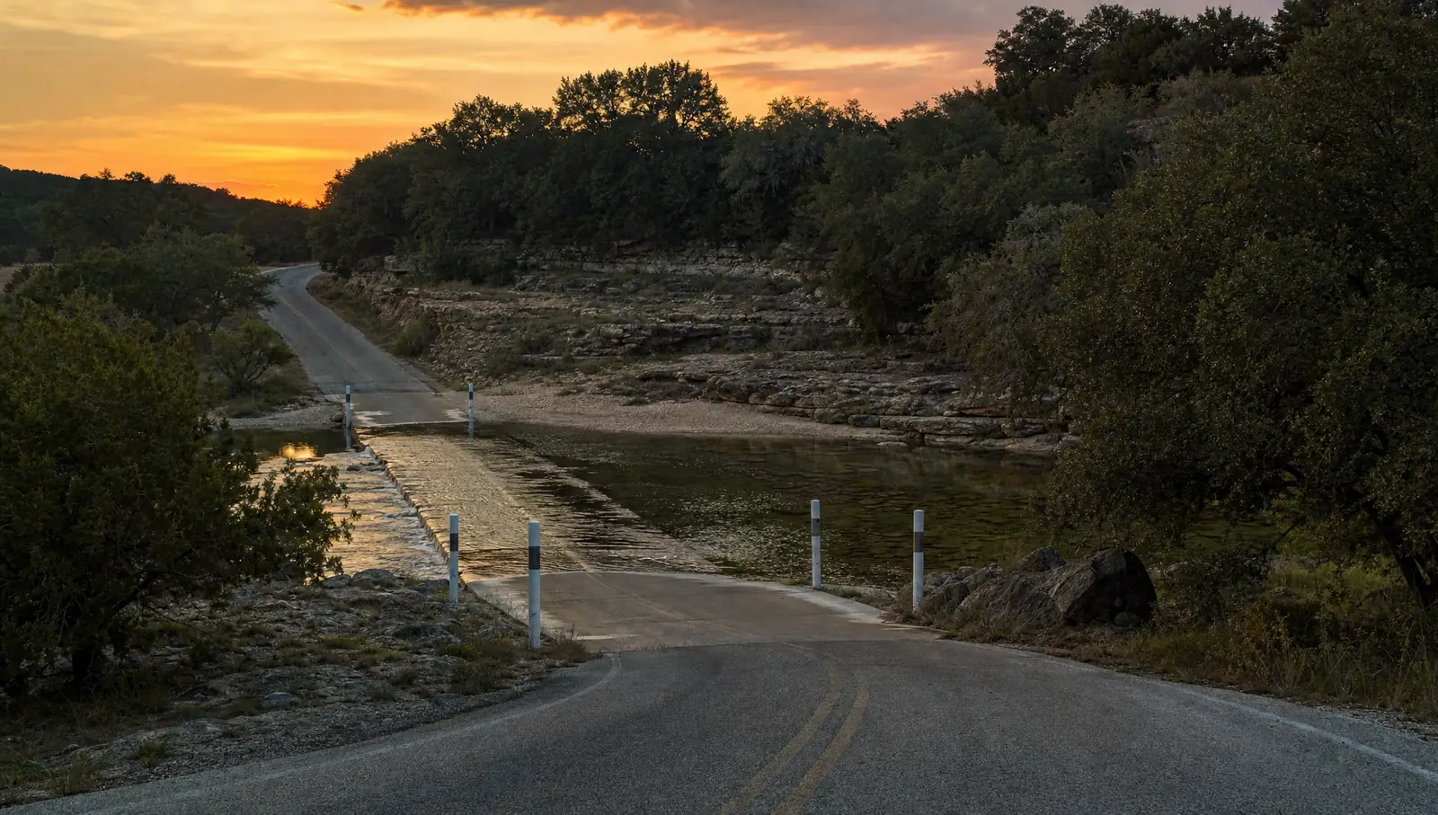 A narrow two-lane road dipping to a low concrete crossing over a shallow limestone creek at dusk, with white marker posts and wooded hills on both sides