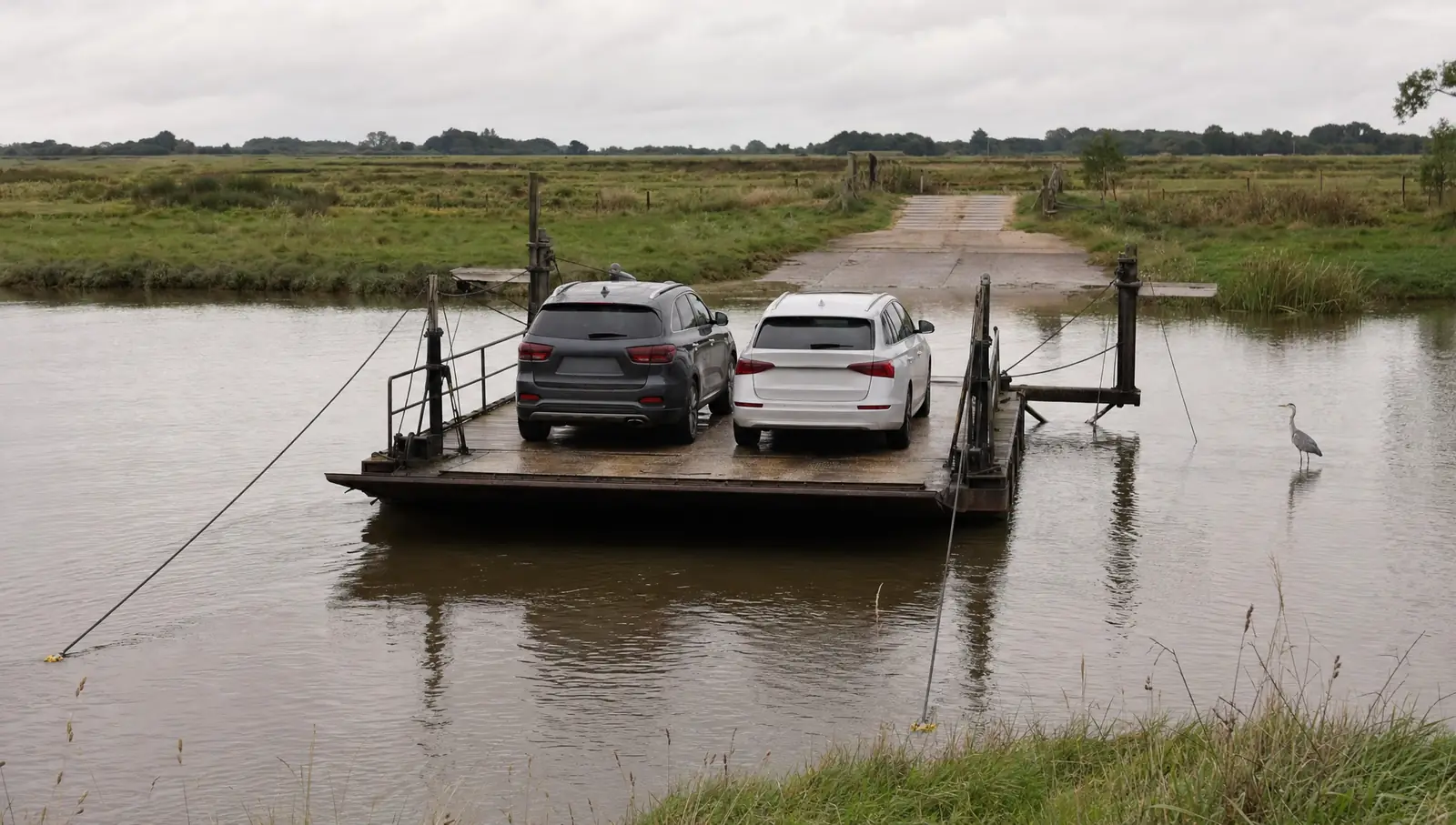 A small flat cable ferry carrying two cars across a narrow tidal river between marsh banks on an overcast morning