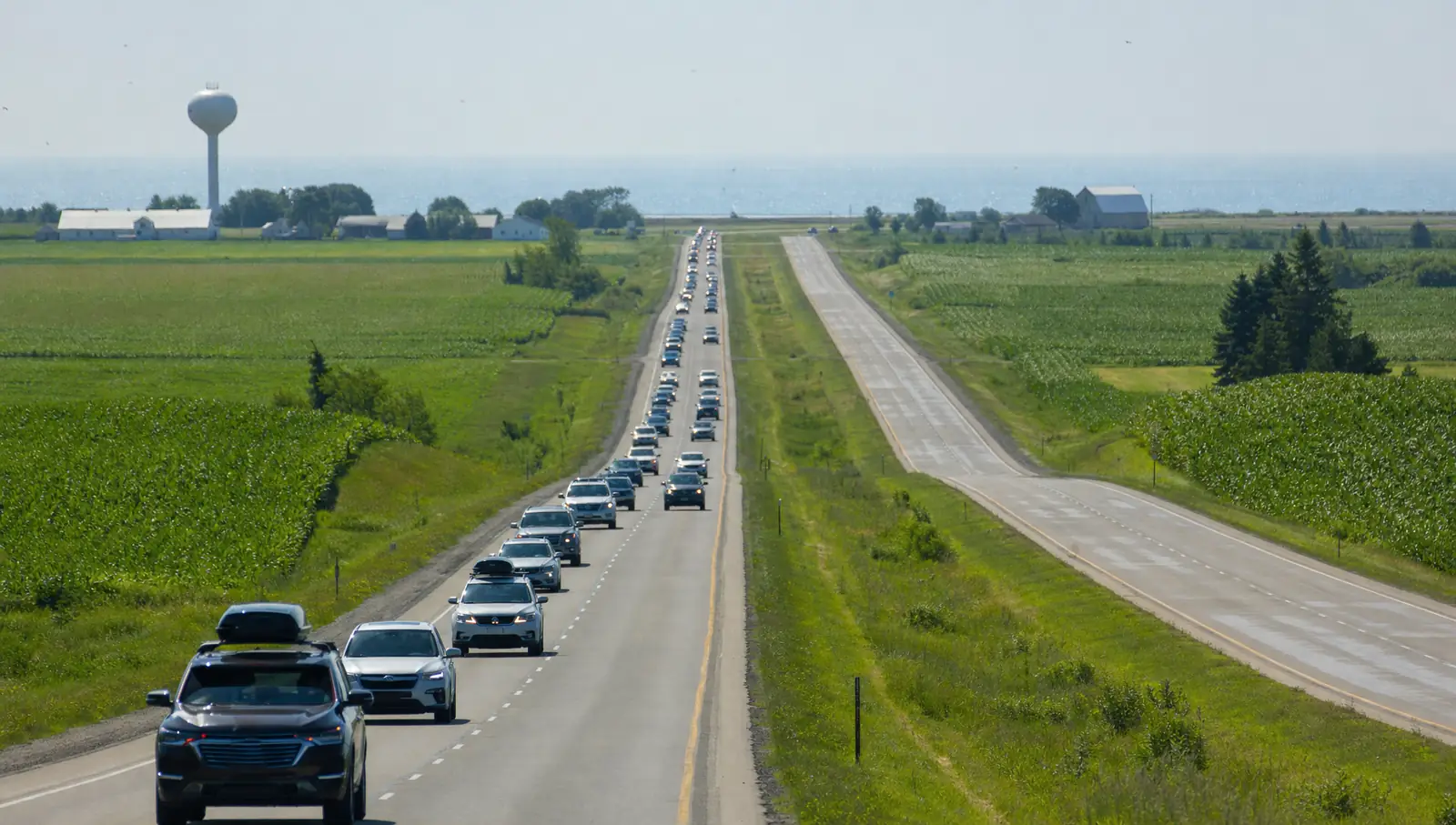 A long line of eastbound cars with roof boxes on a straight divided highway through flat cornfields on a hazy summer afternoon