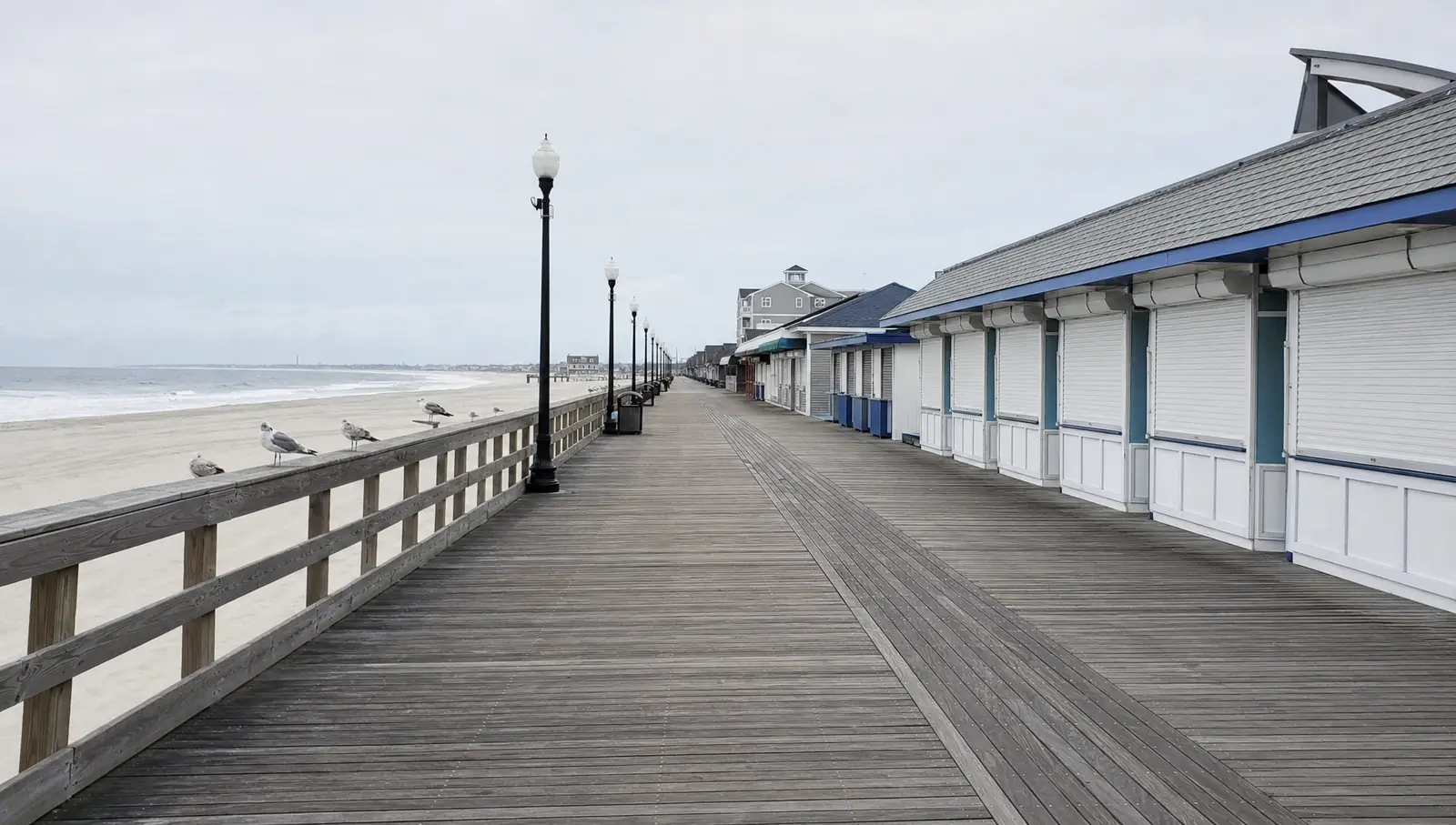An empty wooden boardwalk in a beach town in winter with shuttered stalls, a grey ocean and gulls on the railing