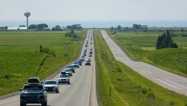 A long line of eastbound cars with roof boxes on a straight divided highway through flat cornfields on a hazy summer afternoon